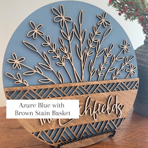 May include: Round wooden sign with an azure blue background and a laser-cut floral design. The bottom section is a brown stain basket with the text "Richfields". The sign is displayed on a black metal stand.