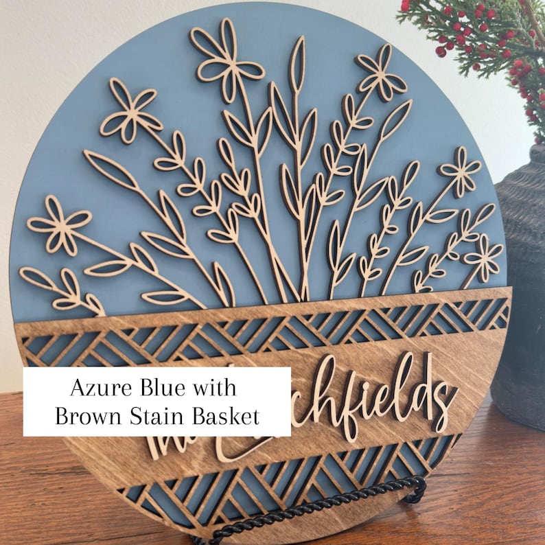 May include: Round wooden sign with an azure blue background and a laser-cut floral design. The bottom section is a brown stain basket with the text "Richfields". The sign is displayed on a black metal stand.