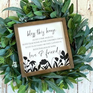 May include: A white wooden sign with black text that reads "Bless this house as we come and go, bless our home as the children grow, bless our family as they gather in, bless this house with love & friends". The sign is decorated with black silhouettes of flowers.