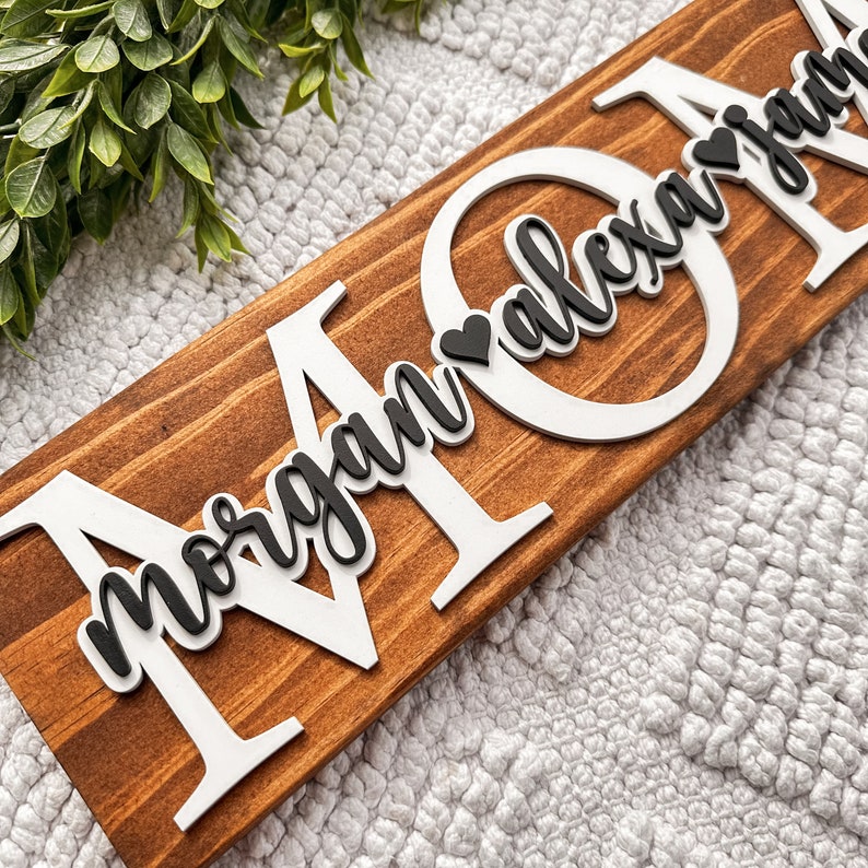 May include: A wooden sign with the word "MOM" in large white letters. The letters are raised and have a heart shape cut out of them. The names "Morgan" and "Alexandra" are written in black cursive letters inside the hearts.