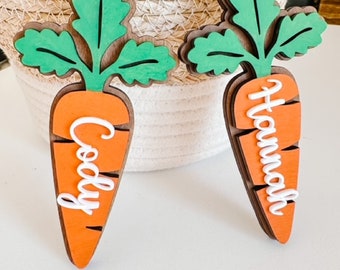 May include: Two wooden carrot ornaments with green tops and the names "Cody" and "Hannah" written in white on the orange carrot bodies.