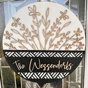 May include: Round wooden sign with a white background and laser-cut floral design. The bottom section is black with a geometric pattern and the family name "The Wessendorfs" in a cursive font. A decorative piece for the home.
