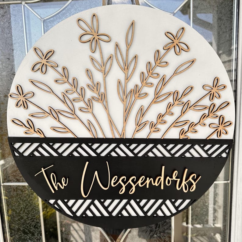 May include: Round wooden sign with a white background and laser-cut floral design. The bottom section is black with a geometric pattern and the family name "The Wessendorfs" in a cursive font. A decorative piece for the home.
