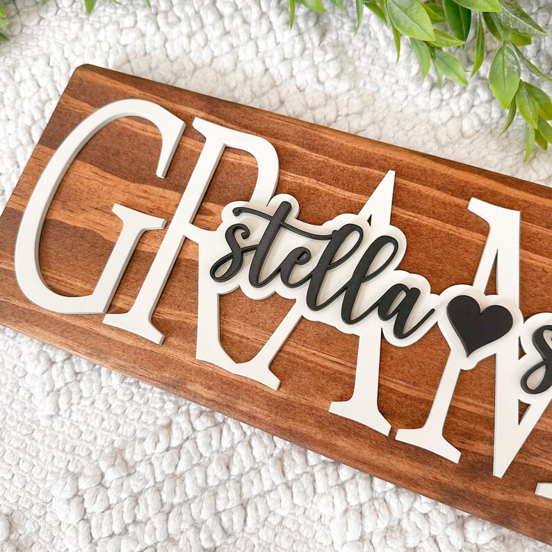 May include: A wooden sign with the word "GRAM" in white letters. The letters are raised and have a 3D effect. The word "stella" is written in black letters on a white background and is placed in front of the word "GRAM". A black heart is placed to the right of the word "stella".