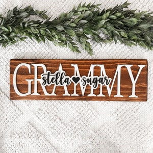 May include: A wooden sign with the word "GRAMMY" in white letters. The letters are raised and have a distressed look. The sign also has the words "stella sugar" in black letters with a small black heart between them.