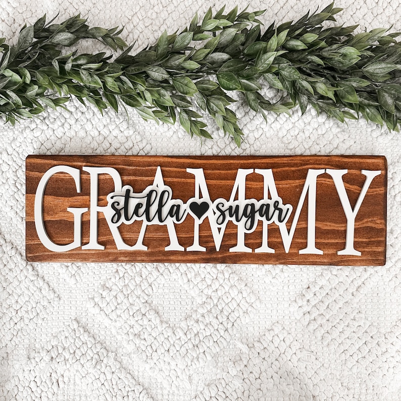 May include: A wooden sign with the word "GRAMMY" in white letters. The letters are raised and have a distressed look. The sign also has the words "stella sugar" in black letters with a small black heart between them.