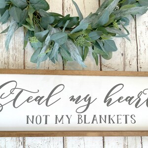 May include: A white wooden sign with a brown frame reads "Steal my heart not my blankets" in gray cursive lettering. A green eucalyptus wreath is above the sign.