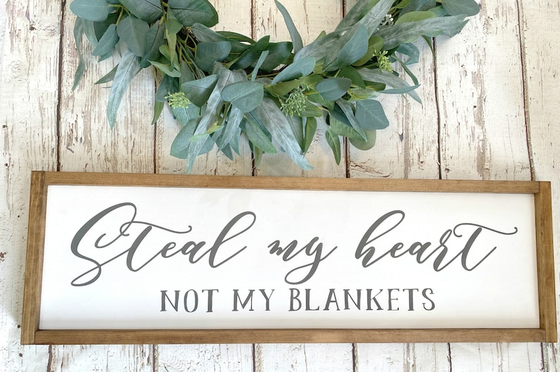 May include: A white wooden sign with a brown frame reads "Steal my heart not my blankets" in gray cursive lettering. A green eucalyptus wreath is above the sign.