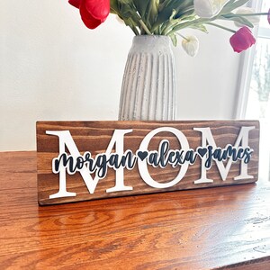 May include: Wooden sign with the word "MOM" in white letters and the names "Morgan", "Alexa", and "James" in black letters with a heart between each name.