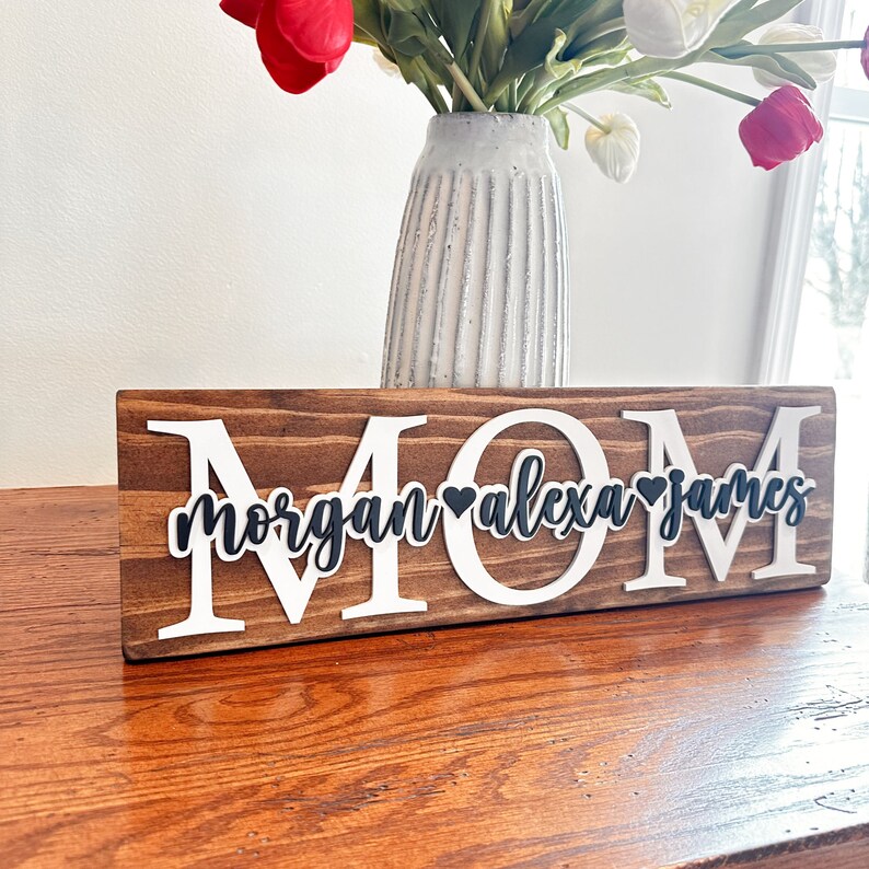 May include: Wooden sign with the word "MOM" in white letters and the names "Morgan", "Alexa", and "James" in black letters with a heart between each name.