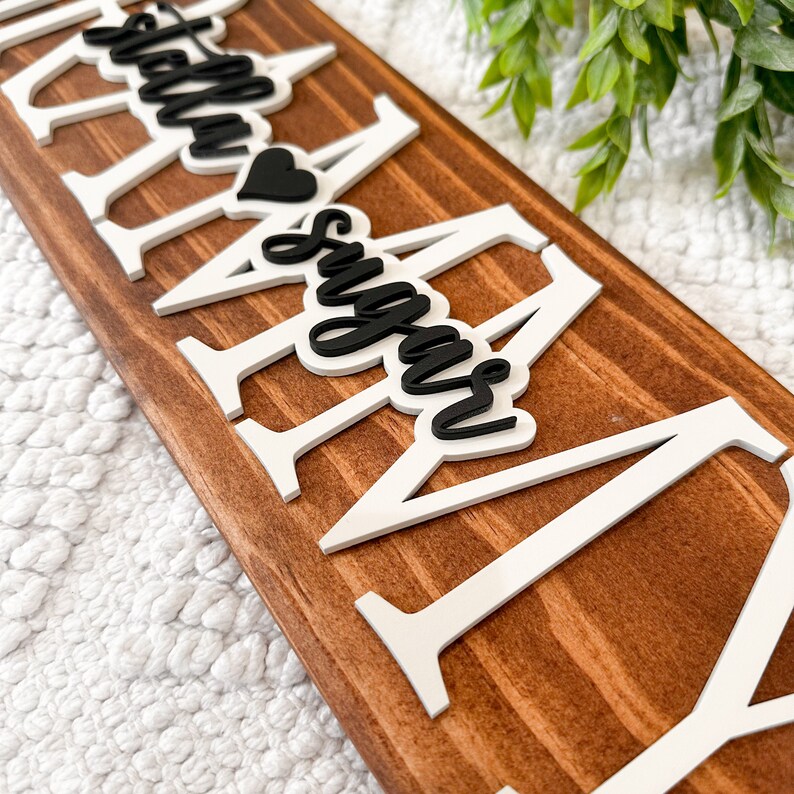 May include: A wooden sign with the words "Stella & Sugar" in black lettering with a heart symbol between the names. The letters are raised and white.