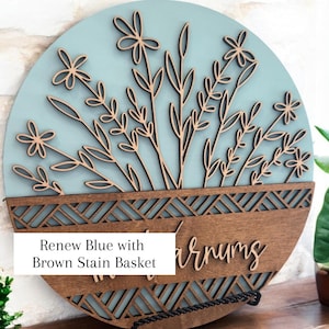 May include: Round wooden wall art with a light blue background and a brown stain basket design. The art features laser-cut flowers and leaves. The text "Renew Blue with Brown Stain Basket" is visible.
