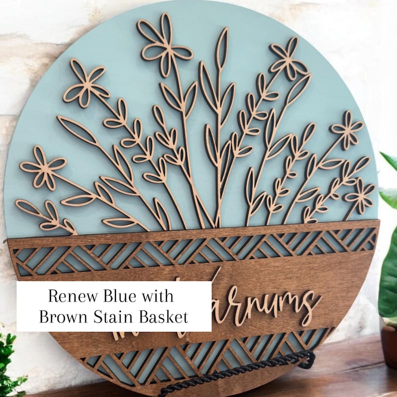 May include: Round wooden wall art with a light blue background and a brown stain basket design. The art features laser-cut flowers and leaves. The text "Renew Blue with Brown Stain Basket" is visible.