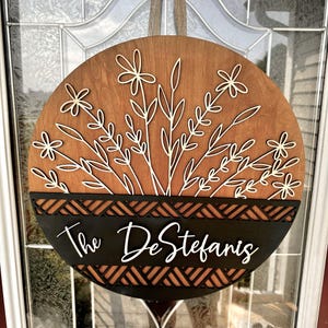 May include: A round wooden door sign with a floral design and the name "The DeStefanis". The sign features a brown background with white floral cutouts and a black lower section with the family name in white script. A decorative piece for the front door.