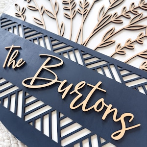 May include: A wooden family name sign with the text "The Burtons" in a cursive font. The sign features a navy blue background with a geometric cut-out design and laser-cut floral accents in a natural wood tone.