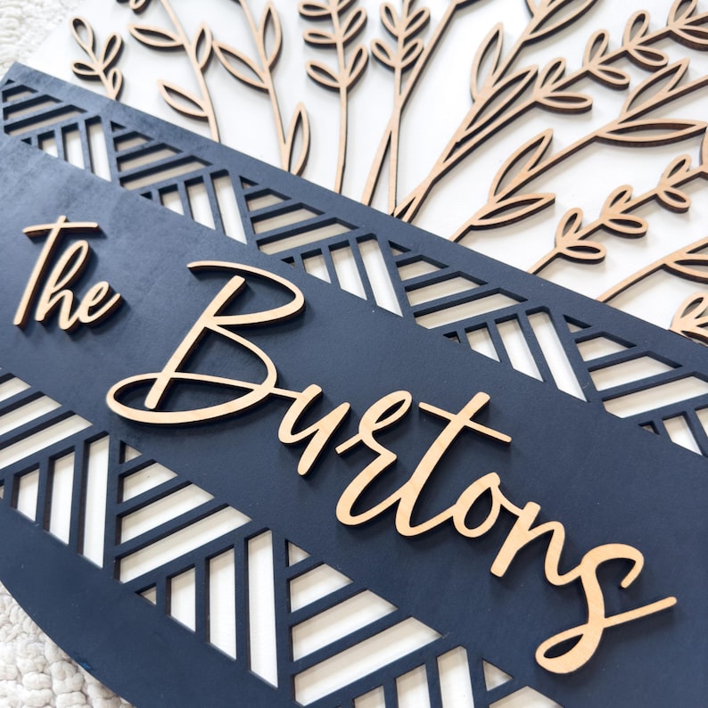 May include: A wooden family name sign with the text "The Burtons" in a cursive font. The sign features a navy blue background with a geometric cut-out design and laser-cut floral accents in a natural wood tone.
