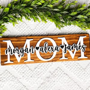 May include: Wooden sign with the word "MOM" in large white letters. The names "Morgan", "Alexa", and "James" are written in black cursive letters with a black heart between each name.