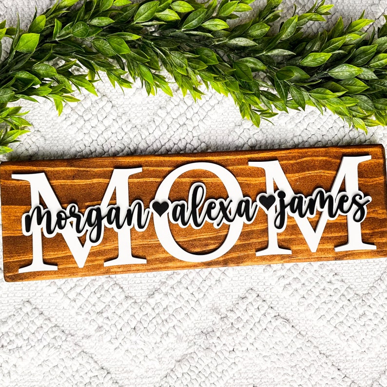 May include: Wooden sign with the word "MOM" in large white letters. The names "Morgan", "Alexa", and "James" are written in black cursive letters with a black heart between each name.