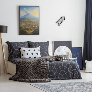 May include: A bedroom with a bed featuring a black and white geometric patterned duvet cover and a gray knitted throw blanket. The bed has a black and white patterned pillow and a white pillow with black crosses. There is a framed landscape painting above the bed, a black arrow wall decor, and a black and white moon print on the wall.