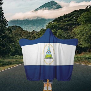 May include: A hooded blanket featuring the flag of Nicaragua. The flag is blue, white, and blue with a triangle in the center. The triangle has a white border and is filled with a blue, white, and green design. The text "REPUBLICA DE NICARAGUA AMERICA CENTRAL" is written around the triangle.