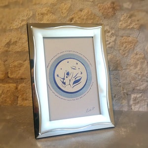 May include: A silver-toned frame with a wavy border holds a print with a blue and white illustration of a person with outstretched arms and a circle of stars. The print includes Hebrew text and the signature "2.16.20".