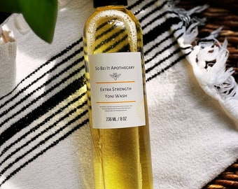 May include: A clear glass bottle with a yellow liquid inside. The bottle is labeled "So Be It Apothecary Extra Strength Yoni Wash 236 mL / 8 oz".