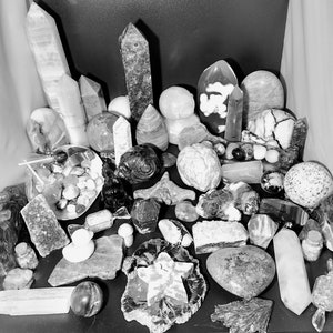 May include: A collection of various crystals and gemstones in shades of white, gray, and black. The assortment includes polished spheres, obelisks, and raw, textured stones. Some crystals are clear, while others have intricate patterns and inclusions. The arrangement is on a dark surface.
