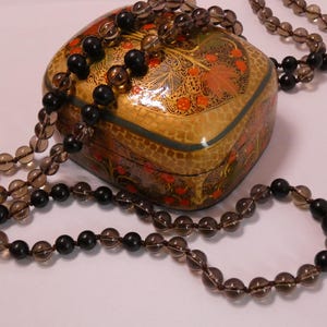May include: A heart-shaped, gold-colored decorative box with intricate floral designs in red, orange, and green. Two necklaces with alternating black and smoky quartz beads are draped over the box.