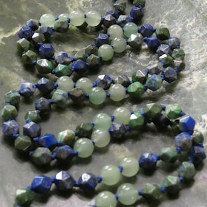 May include: A necklace featuring alternating beads of light green, faceted dark green, and faceted dark blue stones. The beads are strung on a dark blue cord, creating a layered effect. The necklace is displayed on a textured, gray surface.