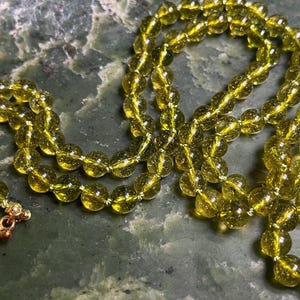 May include: A necklace composed of translucent, olive-green spherical beads with a crackled texture. The strand is secured with a metallic copper-toned toggle clasp featuring a circular ring and a T-bar connector, showing a wire-wrapped design.