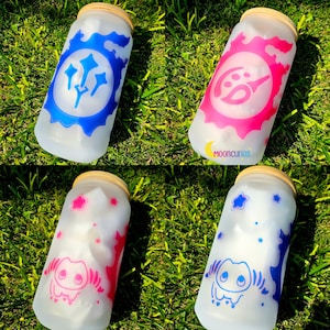 May include: Four frosted glass tumblers with wooden lids are arranged on green grass. Each tumbler features a different design in blue, pink, and white. The text "Mooncurios" is visible on one tumbler. The designs include stars, a sun, and other graphic elements.