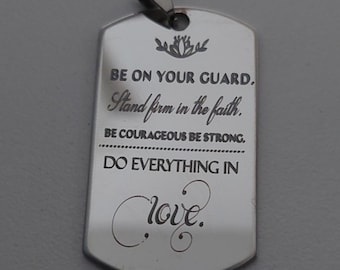 Könnte beinhalten: Ein silberfarbener Anhänger in Form einer Erkennungsmarke mit einer Schlaufe oben. Der Anhänger enthält eingravierten Text, darunter "BE ON YOUR GUARD, Stand firm in the faith, BE COURAGEOUS BE STRONG, DO EVERYTHING IN love."