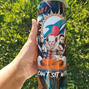 May include: A stainless steel tumbler featuring a design with the Miami Dolphins logo, horror movie characters, and the text "CAN'T SIT WITH". The tumbler has a black and orange background with a silver lid.