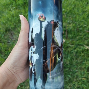May include: A tall, cylindrical tumbler featuring a spooky Halloween design. The artwork depicts the Headless Horseman riding a horse, holding a jack-o'-lantern, set against a dark, moonlit sky. The tumbler has a silver lid and is held by a hand.