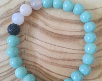 May include: A turquoise and white beaded bracelet with a black lava bead. The bracelet is made of round beads and is on a wooden surface.