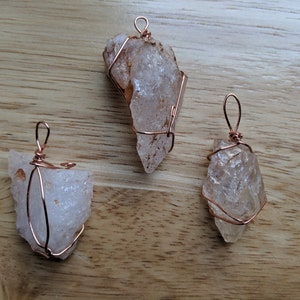 May include: Three clear quartz crystal pendants wrapped in copper wire. The crystals are all different shapes and sizes. The wire is a rose gold color.