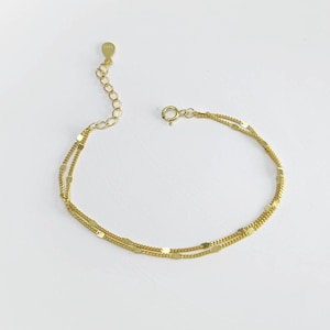 Könnte beinhalten: Ein zartes goldenes Armband mit zwei Strängen aus kleinen, flachen, rechteckigen Gliedern. Das Armband hat eine verstellbare Kette und einen kleinen Verschluss. Der Schmuck wird auf einer weißen Oberfläche präsentiert.