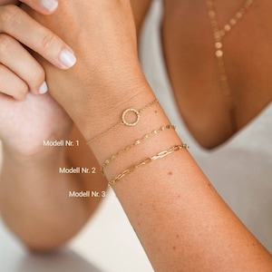 Peut inclure: Trois bracelets en chaîne dorée sur le poignet d'une personne. Le bracelet du haut a un pendentif rond. Les bracelets sont étiquetés "Modell Nr. 1", "Modell Nr. 2" et "Modell Nr. 3".