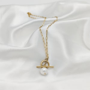 Könnte beinhalten: Eine goldene Kettenkette mit einem Toggle-Verschluss und einem einzelnen Perlenanhänger.