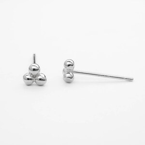 Può includere: Un paio di orecchini a lobo in argento. Ogni orecchino presenta tre piccole perle rotonde e lucide in argento raggruppate. Gli orecchini sono esposti su uno sfondo bianco, mostrando il loro design minimalista e la superficie riflettente.
