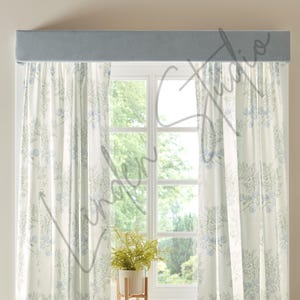 May include: A window with sheer white curtains featuring a blue floral pattern. A light blue valance sits above the window. A small potted plant sits on a wooden stand in front of the window. A lamp and chair are in the foreground.