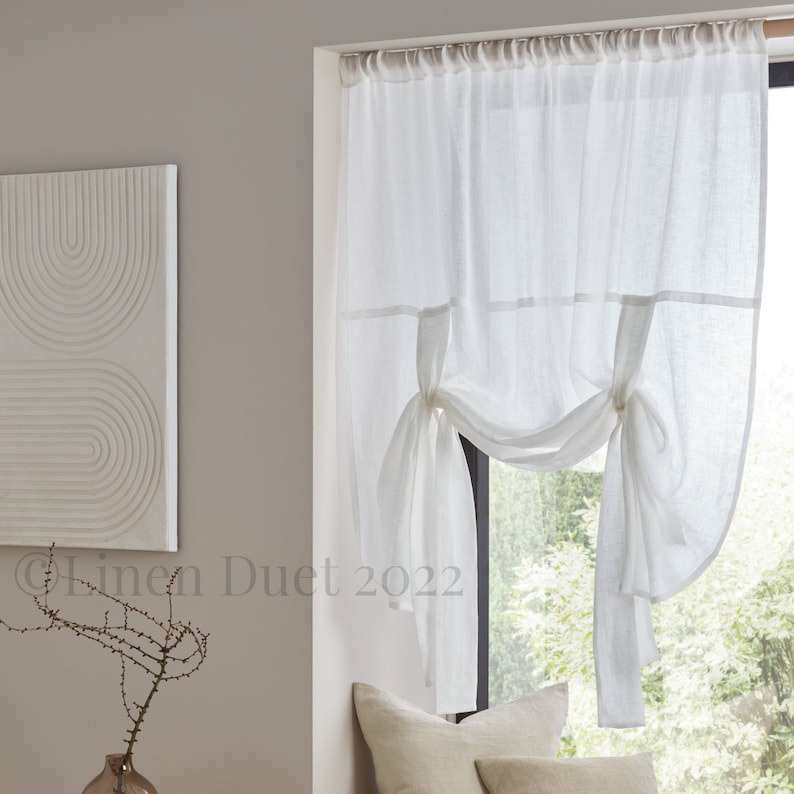 May include: White sheer linen curtains with a gathered design and tied back with fabric ties. The curtains are hung on a window with a view of green trees.