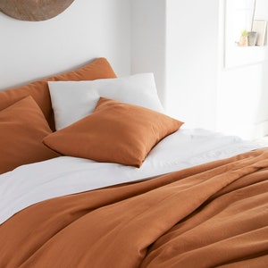 Puede incluir: Una funda n&oacute;rdica de lino de color &oacute;xido con una s&aacute;bana de lino blanca y dos almohadas de lino de color &oacute;xido en una cama.