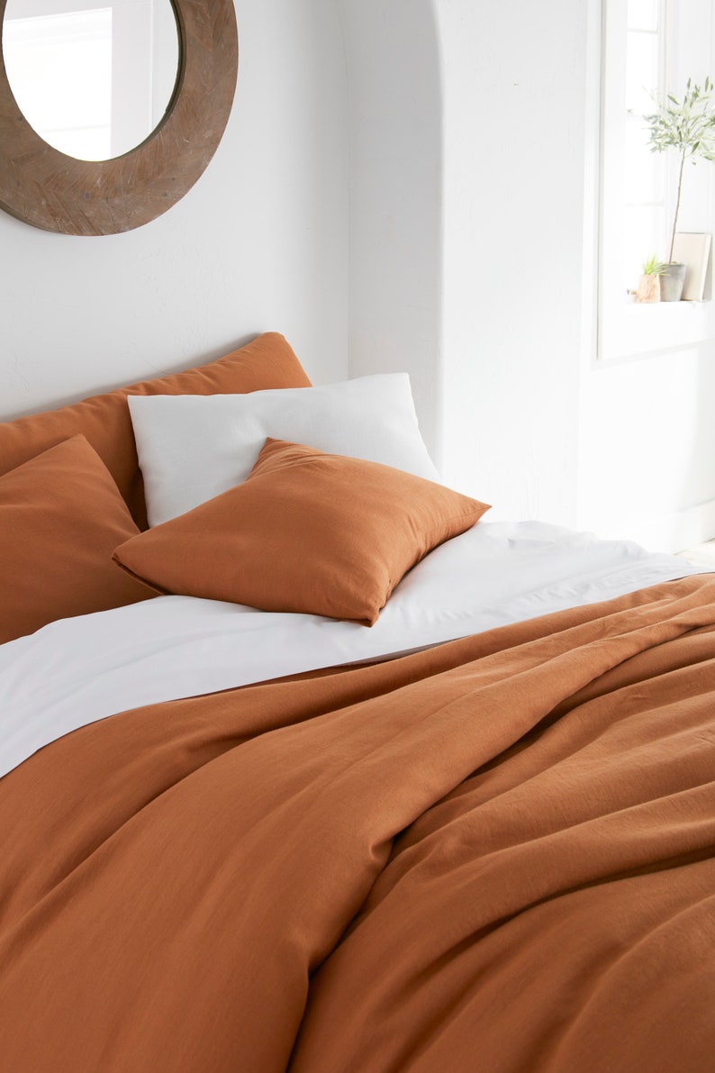 Puede incluir: Una funda n&oacute;rdica de lino de color &oacute;xido con una s&aacute;bana de lino blanca y dos almohadas de lino de color &oacute;xido en una cama.