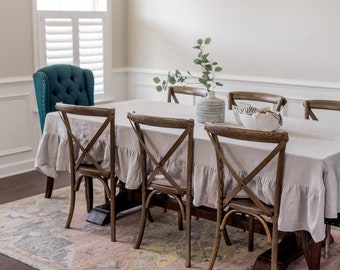 May include: A dining room scene featuring a wooden table set with a light gray linen tablecloth with a ruffled edge. Wooden chairs with an X-shaped back surround the table. A teal upholstered chair is at the head of the table. A vase with greenery sits on the table.