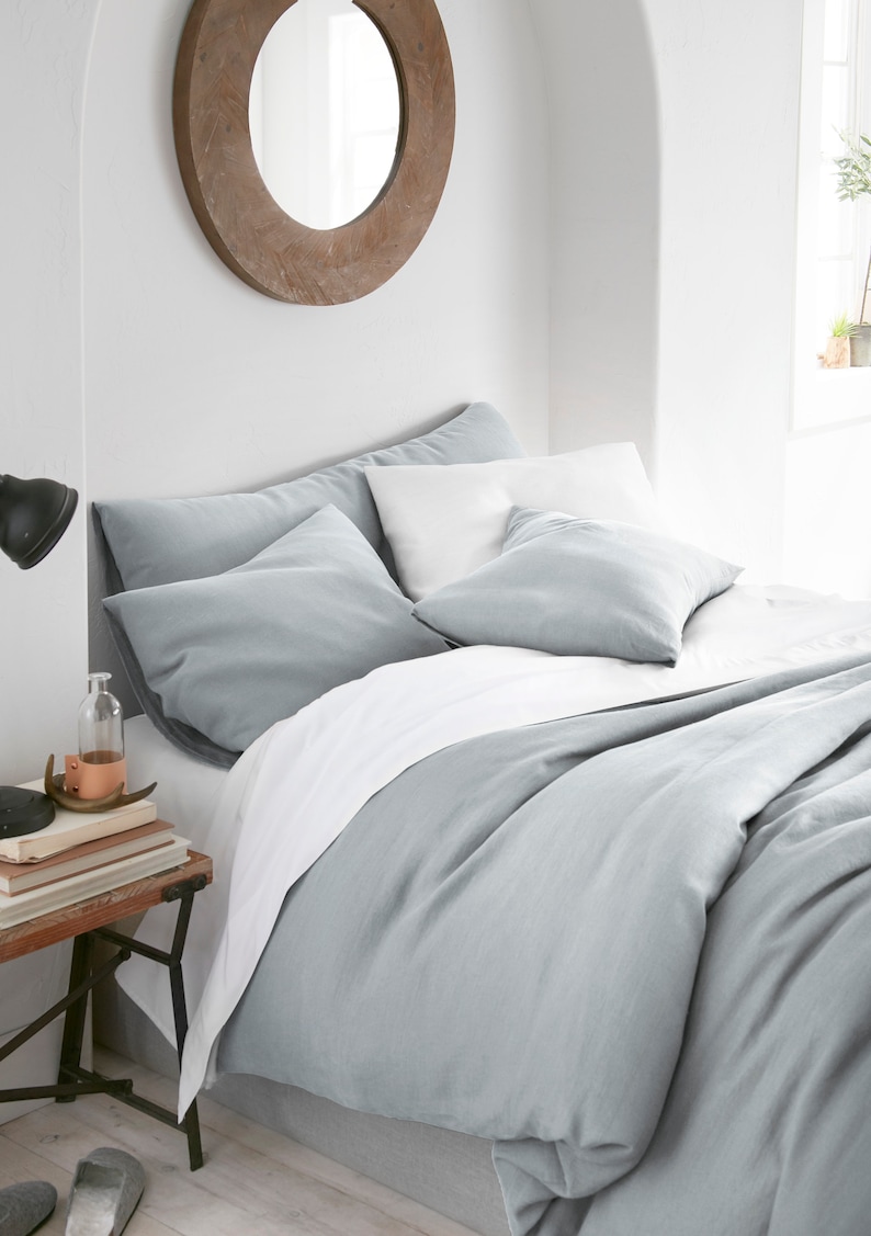 Puede incluir: Una funda n&oacute;rdica de lino azul claro con una s&aacute;bana de lino blanco y dos fundas de almohada de lino azul claro. La ropa de cama est&aacute; en una cama con una cabecera de madera y un espejo redondo encima de la cama.