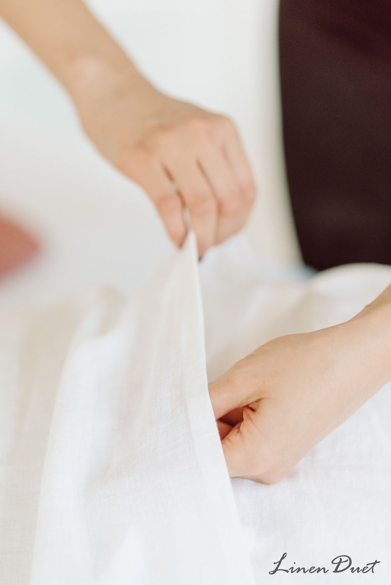 Puede incluir: Una persona haciendo la cama con s&aacute;banas de lino blanco. Las s&aacute;banas son lisas y sin arrugas. Las manos de la persona son visibles mientras mete las s&aacute;banas en el colch&oacute;n.