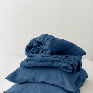May include: A stack of blue linen bedding, including a fitted sheet, flat sheet, and pillowcases. The bedding is folded neatly and arranged on a white surface.