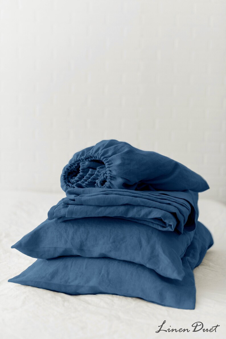 May include: A stack of blue linen bedding, including a fitted sheet, flat sheet, and pillowcases. The bedding is folded neatly and arranged on a white surface.