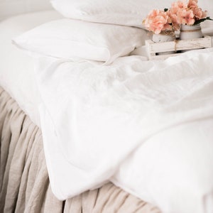 May include: White linen bedding with a tan linen bed skirt.  A small wooden tray with pink flowers in glass jars sits on the bed.